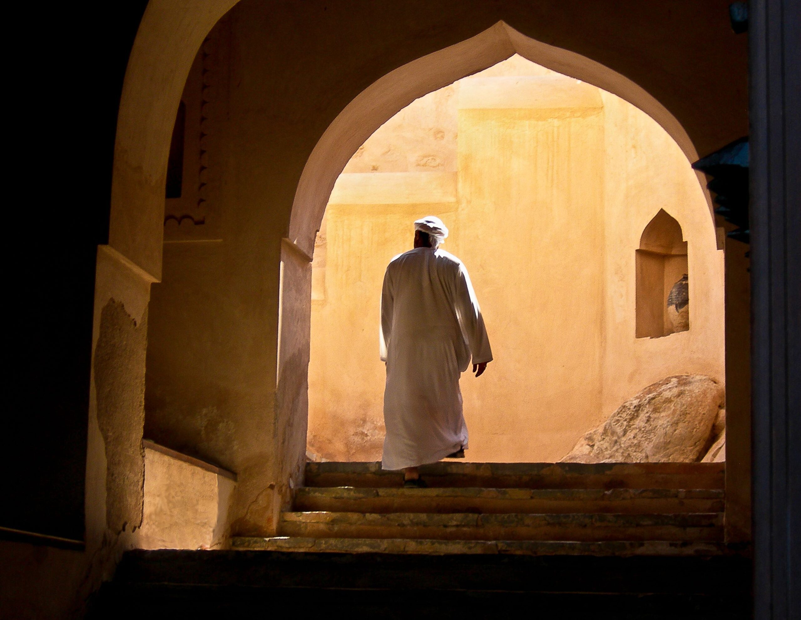 Uomo in abito tradizionale omanita che cammina sotto un arco illuminato. Viaggi Oman Monica Alberghini.