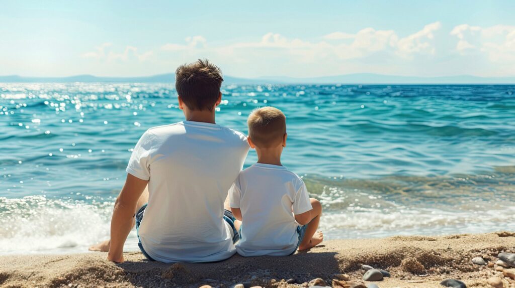 Papà single e figlio seduti insieme su una spiaggia, guardando il mare, simbolo di viaggi indimenticabili e proposte di viaggio per famiglie di papà single, a cura di Monica Alberghini.
