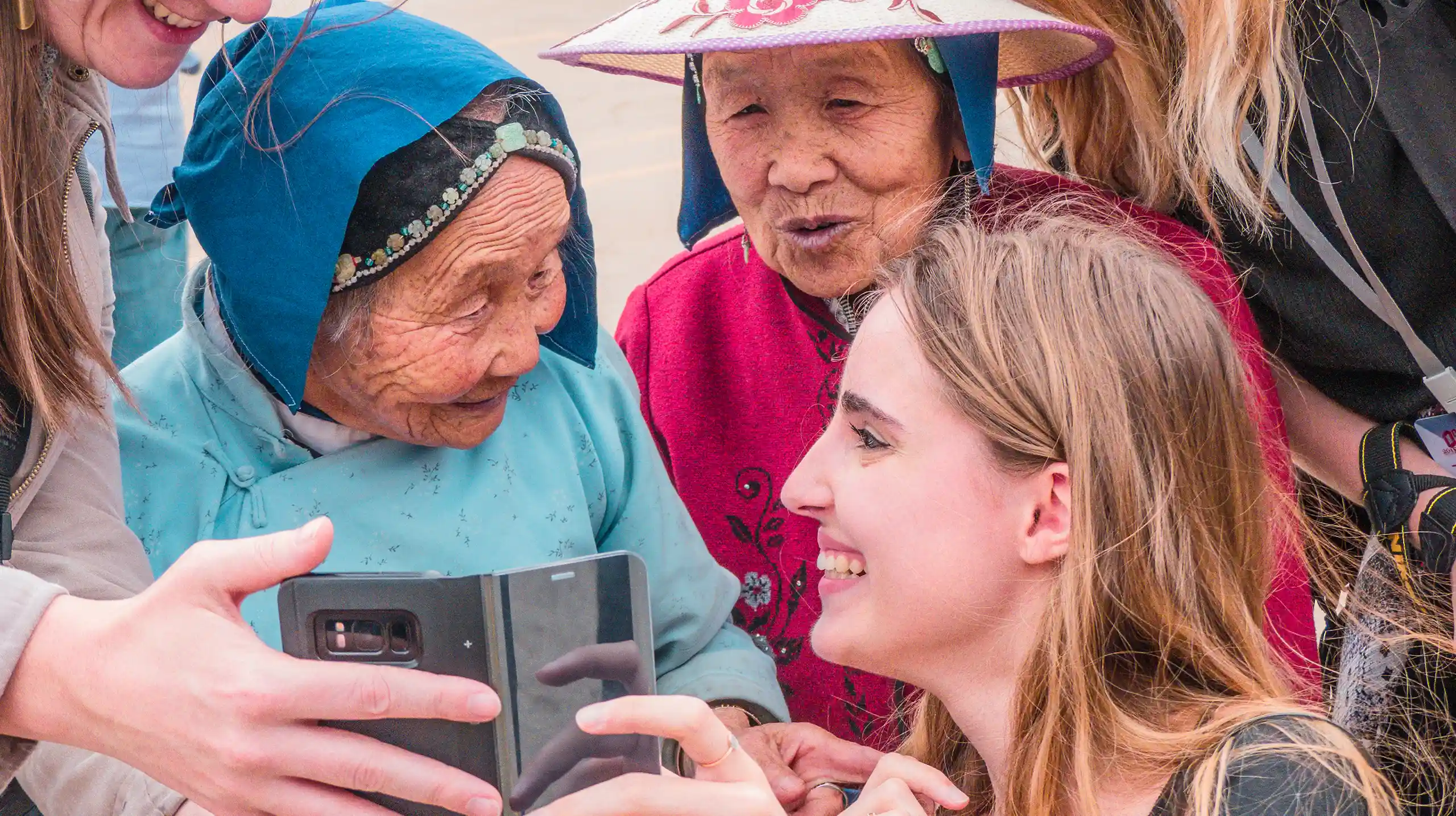 Giovani viaggiatori condividono un momento con donne anziane locali, guardando un telefono insieme e sorridendo. Viaggi esperienziali Monica Alberghini