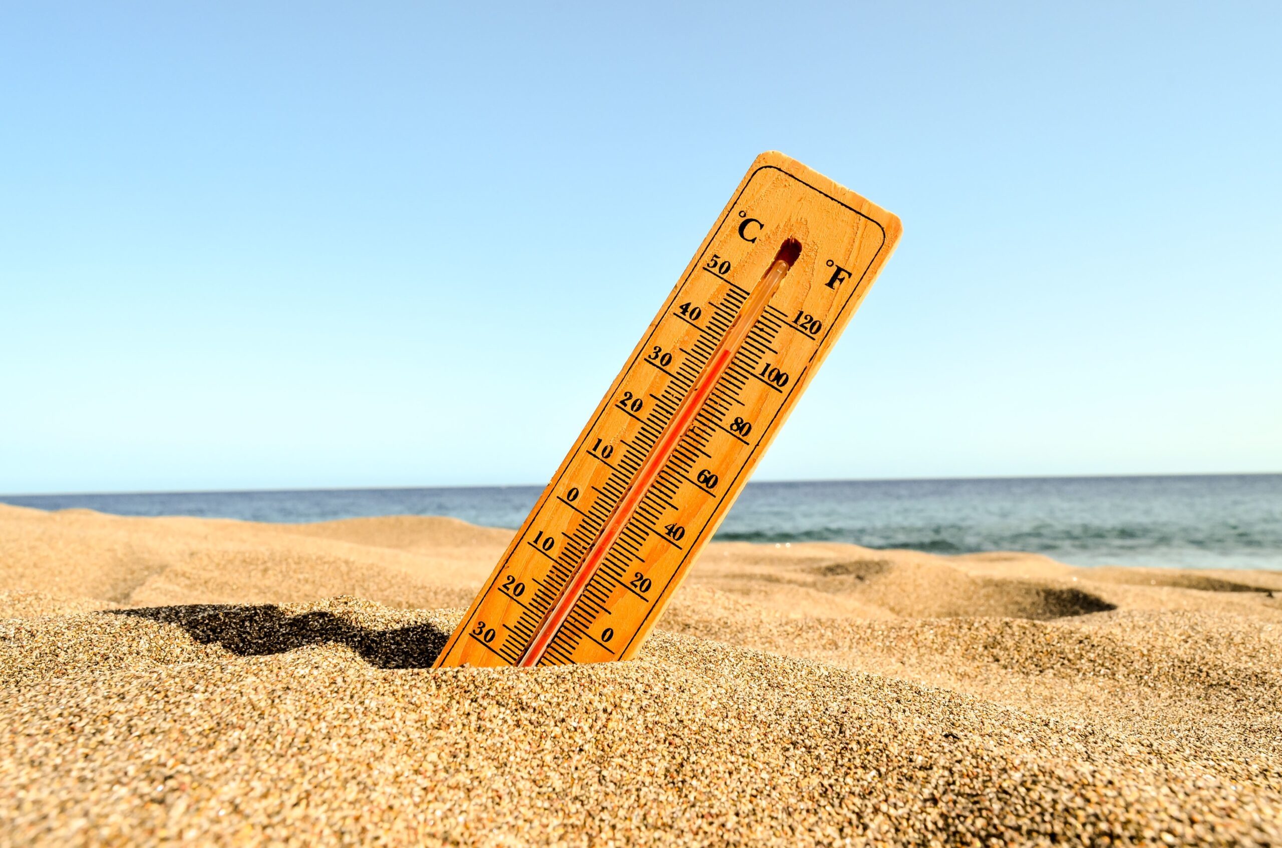 Termometro infilato nella sabbia su una spiaggia assolata, con il mare sullo sfondo. L'immagine rappresenta l'importanza di considerare il clima nella pianificazione di un viaggio per evitare temperature estreme e scegliere il momento ideale per partire.