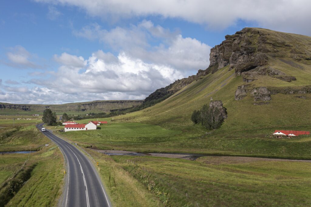 Strada panoramica che attraversa un paesaggio verde in Islanda, con fattorie tradizionali ai piedi delle montagne
